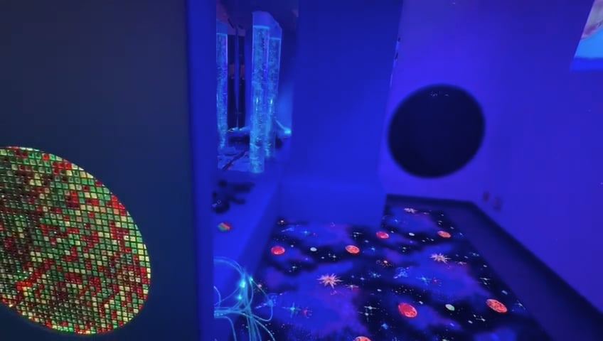 Load video: This is a video showing a sensory room that has soft, purple lighting, and padded furniture. There are bubble tubes with fish inside and projectors projecting and aquarium with fish in it on to the wall.